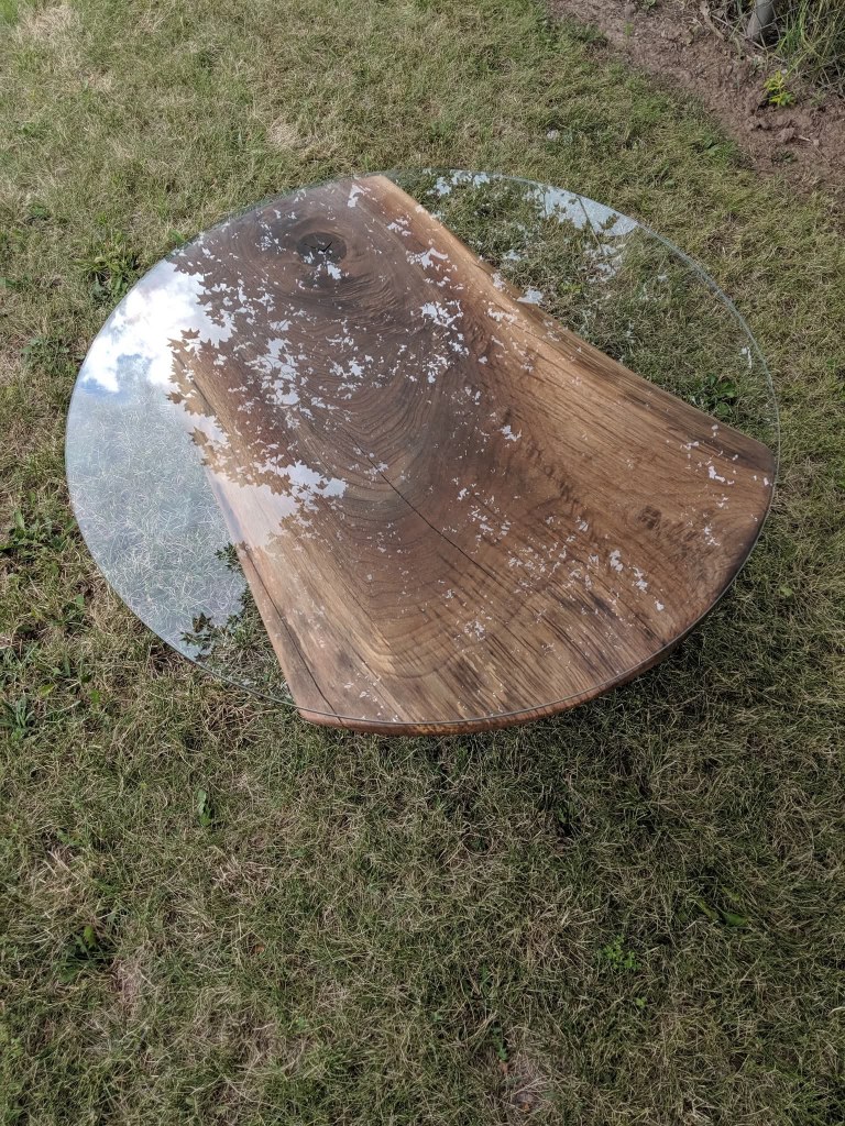 A glass table with a wooden base, placed on grass, with a reflection of the sky and trees.