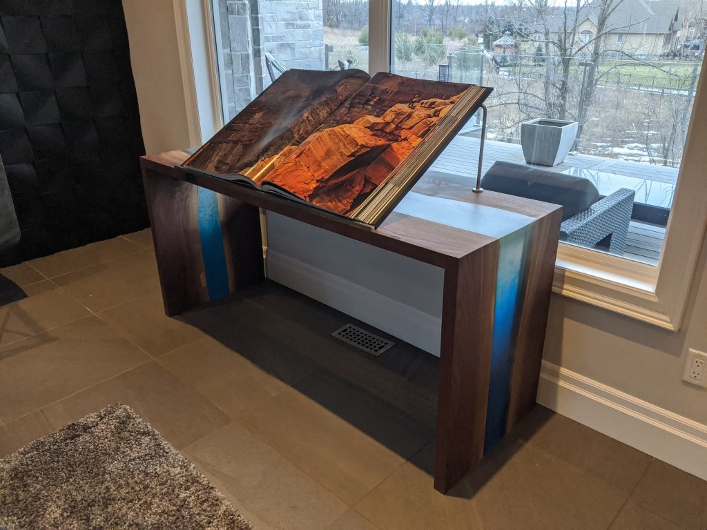 A wooden table with a large book open on it, positioned in front of a window. The book's pages display a landscape image, ...