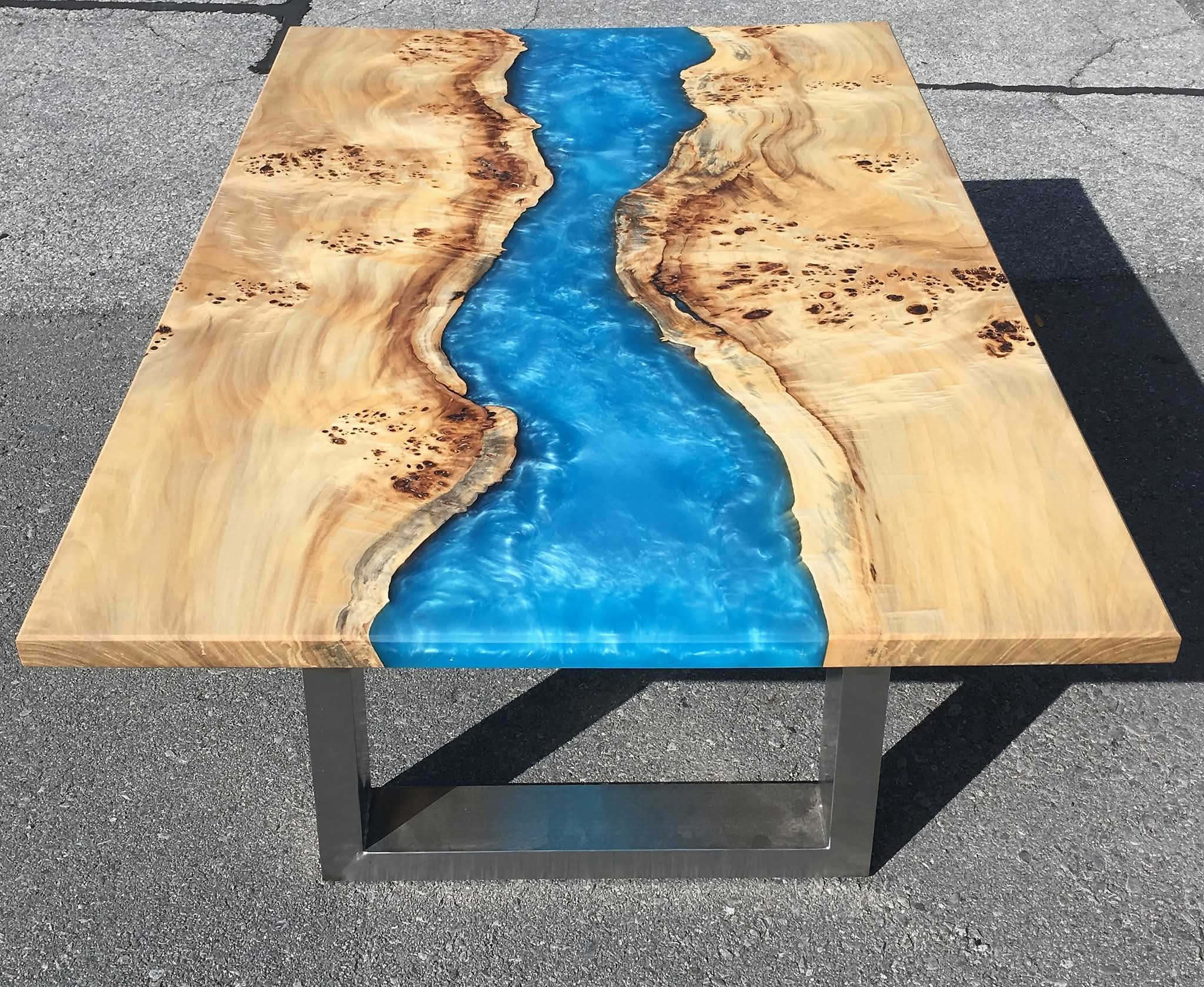 A square table made of wood and resin with a blue river running through it. The wood is light brown with darker brown spot...